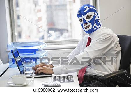 Stock Image - Portrait of businessman using laptop while wearing wrestling mask at office desk. Fotosearch