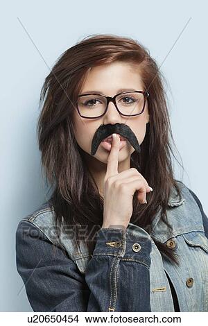 Picture - Portrait of young woman with fake mustache and finger on her lips against light blue background. Fotosearch