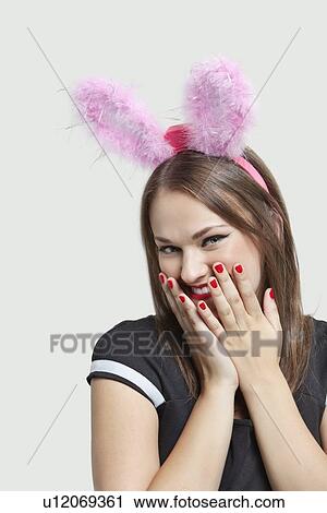 Stock Image - Shy young woman wearing rabbit ears over gray background. Fotosearch