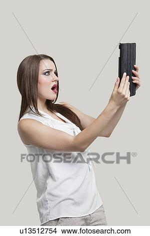 Picture - Side view of a shocked young woman looking at tablet PC over gray background. Fotosearch