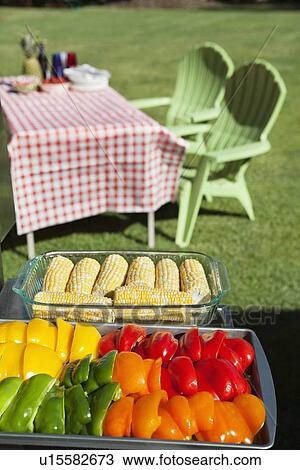 Stock Image - Slices of bell peppers and sweet corn in glass containers with dining table and chairs at lawn. Fotosearch