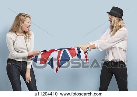 Stock Image - Two young women playing tug of war with British flag against light blue background. Fotosearch