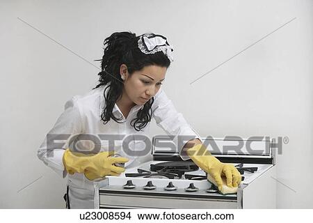 Picture - Young maidservant cleaning stove against gray background. Fotosearch