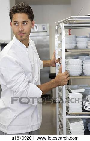 Stock Image - Young male chef by rack of utensils. Fotosearch