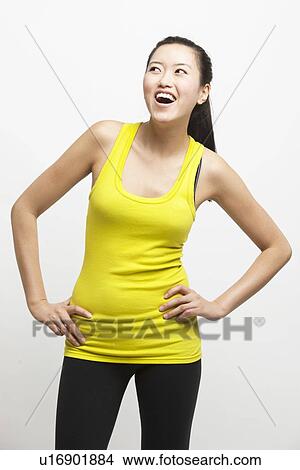 Picture - Happy young woman in yellow tank top with hands on hips looking away against white background. Fotosearch