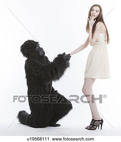 Stock Image - Man in gorilla costume proposing surprised young woman against white background. Fotosearch