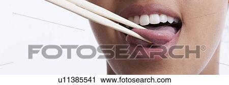 Stock Image - Playful young woman with chopsticks sticking out tongue against white background. Fotosearch