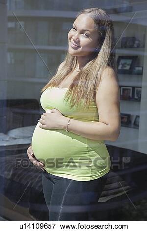 Portrait of a happy pregnant young woman standing with hands on stomach View Large Photo Image Stock Photo - Portrait of a happy pregnant young woman standing with hands on stomach. Fotosearch