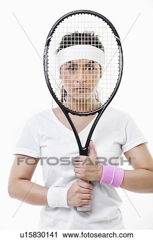 Stock Image - Portrait of an Asian young woman looking through tennis racket over white background. Fotosearch