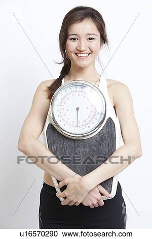 Portrait of cheerful Asian woman holding weight scale against white background View Large Photo Image Stock Image - Portrait of cheerful Asian woman holding weight scale against white background. Fotosearch