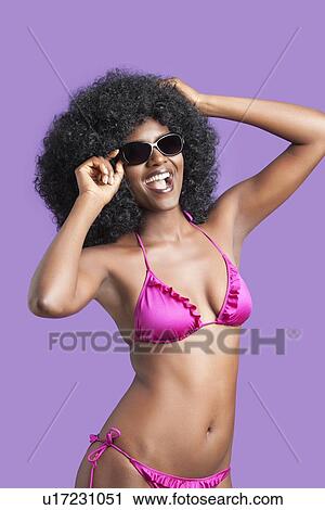 Stock Image - Portrait of happy young woman in sunglasses and bikini posing over violet background. Fotosearch