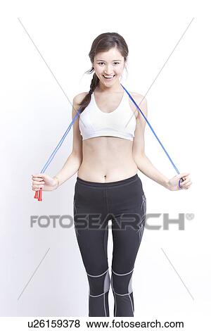 Stock Photo - Portrait of smiling young woman exercising with skipping rope against white background. Fotosearch