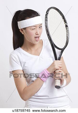 Stock Photography - Portrait of young Asian woman with tennis racket winking against white background. Fotosearch