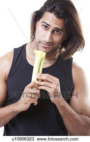 Stock Image - Portrait of young muscular man holding eating celery over white background. Fotosearch
