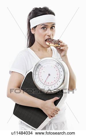 Portrait of young woman eating donut holding weight scale over white background View Large Photo Image Picture - Portrait of young woman eating donut holding weight scale over white background. Fotosearch