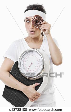 Stock Photo - Portrait of young woman looking through donut holding weight scale over white background. Fotosearch