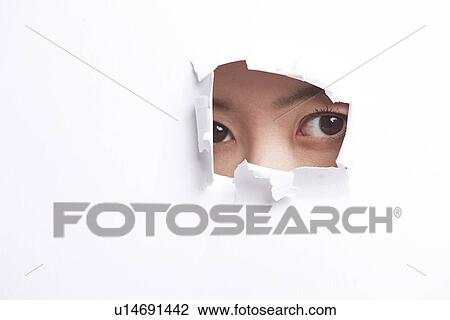 Stock Image - Portrait of young woman peeking through a ripped paper hole. Fotosearch