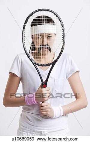 Stock Photo - Portrait of young woman with a fake mustache holding tennis racket in front of her face against white background. Fotosearch