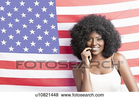 Stock Photo - Portrait of young woman with fizzy hairstyle standing against American flag. Fotosearch