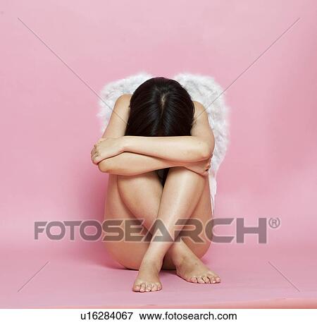 Stock Photo - Sad Asian woman with angel wings sitting on floor against pink background. Fotosearch