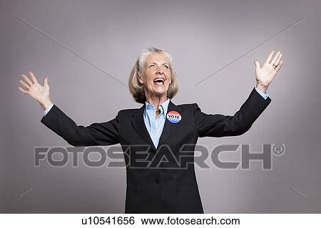 Stock Photograph - Senior businesswoman wearing Vote Badge with arms raised on grey background. Fotosearch