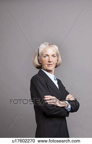 Stock Image - Senior businesswoman with arms folded standing infront of a grey background. Fotosearch