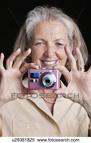 Stock Photography - Smiling senior woman taking picture with camera against black background. Fotosearch