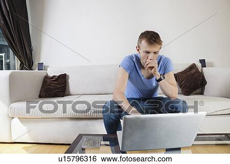 Thoughtful man using laptop while sitting on sofa at home View Large Photo Image Stock Photograph - Thoughtful man using laptop while sitting on sofa at home. Fotosearch