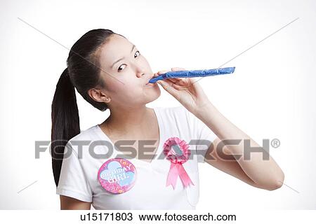 Stock Image - Young Asian woman blowing noisemaker against white background. Fotosearch