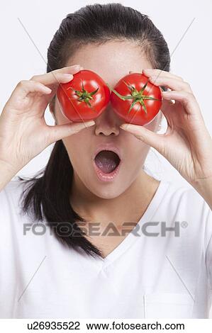Stock Image - Young Asian woman covering her eyes with tomatoes against white background. Fotosearch