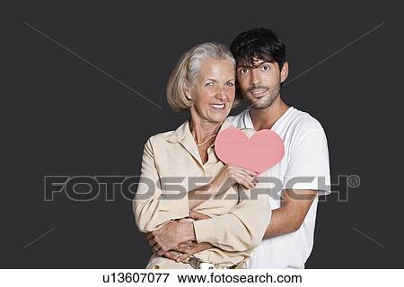 Young man embracing senior woman holding red paper heart against black background View Large Photo Image Stock Photo - Young man embracing senior woman holding red paper heart against black background. Fotosearch