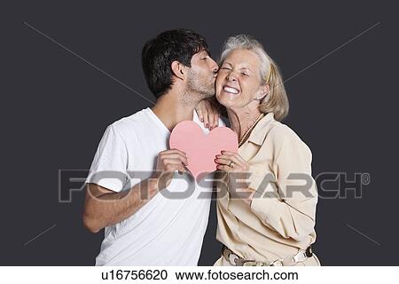 Young man kissing senior woman with red paper heart against black background View Large Photo Image Stock Image - Young man kissing senior woman with red paper heart against black background. Fotosearch