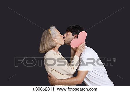 Stock Image - Young man kissing senior woman with red paper heart against black background. Fotosearch