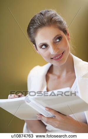 Stock Image - Young woman looking sideways while writing notes on spiral notebook. Fotosearch
