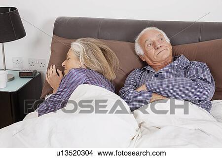 Picture - Bored elderly man looking away while spouse sleeping besides in bedroom. Fotosearch