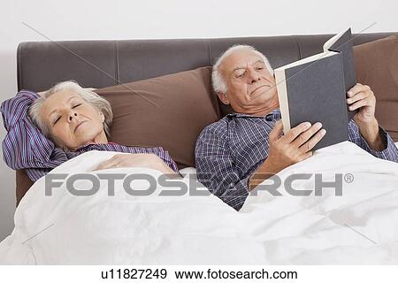 Elderly man reading book while spouse sleeping besides in bedroom View Large Photo Image Stock Photo - Elderly man reading book while spouse sleeping besides in bedroom. Fotosearch