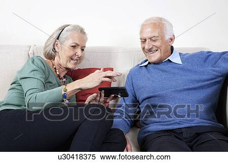 Stock Photography - Happy senior couple using smart phone while sitting on sofa at home. Fotosearch
