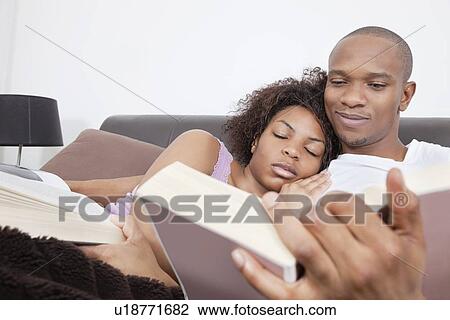 Stock Image - Happy young man reading book as woman sleeping on his shoulder. Fotosearch
