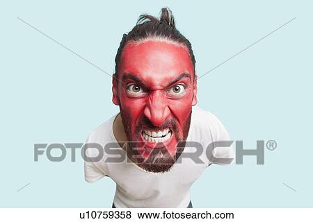 Stock Photo - Portrait of aggressive young man with red painted face clenching teeth against blue background. Fotosearch