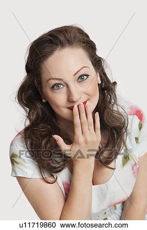 Stock Image - Portrait of beautiful young Caucasian woman smiling with hand over mouth against white background. Fotosearch