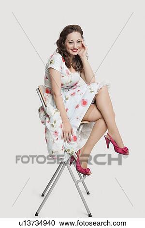 Portrait of beautiful young woman in dress sitting on stool against white background View Large Photo Image Stock Image - Portrait of beautiful young woman in dress sitting on stool against white background. Fotosearch
