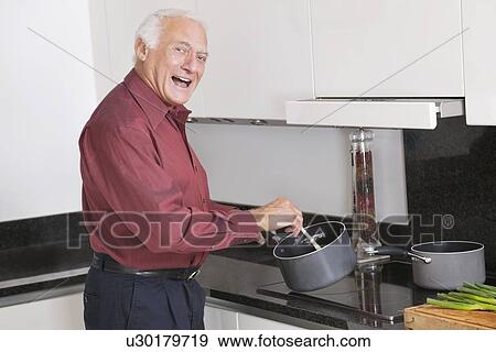 Stock Photo - Portrait of cheerful elderly man preparing food in kitchen. Fotosearch