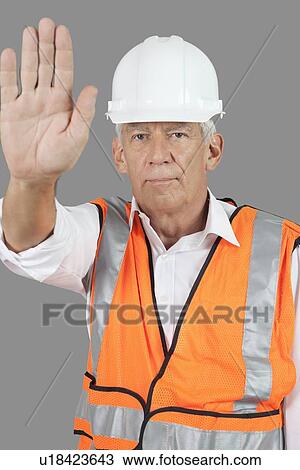 Stock Image - Portrait of confident male construction worker with stop gesture over gray background. Fotosearch