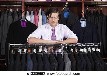 Stock Image - Portrait of confident man leaning on clothing rack in store. Fotosearch