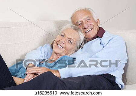 Stock Image - Portrait of happy elderly man with arm around spouse lying on sofa at home. Fotosearch
