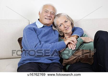 Portrait of happy elderly man with arm around spouse sitting on sofa at home View Large Photo Image Stock Photo - Portrait of happy elderly man with arm around spouse sitting on sofa at home. Fotosearch