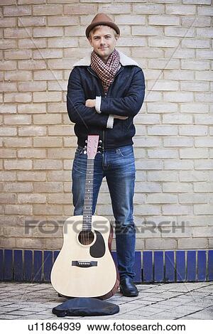 Stock Photo - Portrait of happy young man with guitar standing against brick wall. Fotosearch