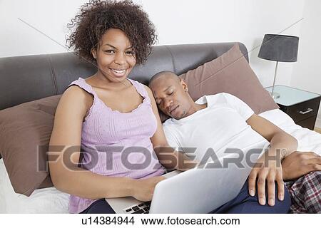 Portrait of happy young woman using laptop while men sleeping in bed View Large Photo Image Picture - Portrait of happy young woman using laptop while men sleeping in bed. Fotosearch