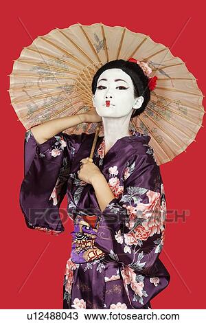 Stock Image - Portrait of Japanese woman in kimono holding parasol against red background. Fotosearch