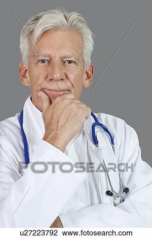 Stock Image - Portrait of senior male doctor with hand on chin over gray background. Fotosearch